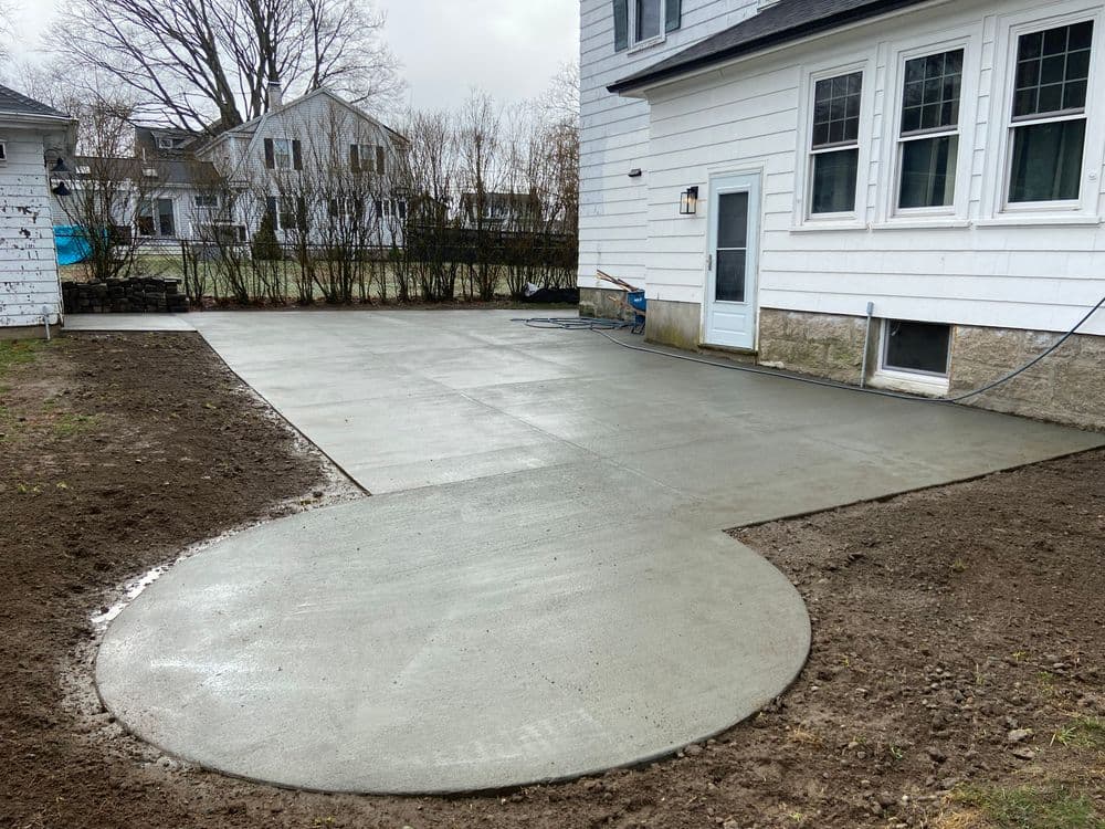 Newly poured concrete patio with circular section next to a white house and grassy area.