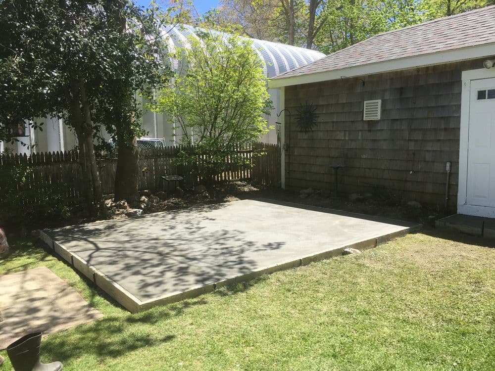 Concrete slab in a backyard, surrounded by trees and a wooden fence, ready for construction.